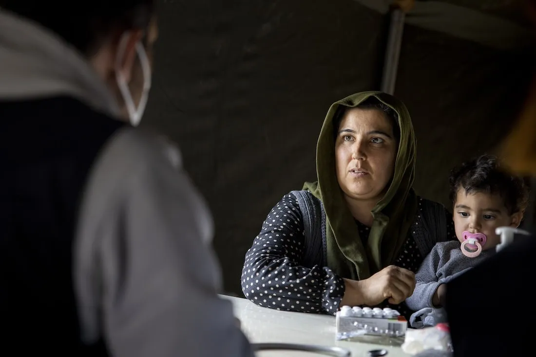 mother holding child speaks to medical staff in tent.