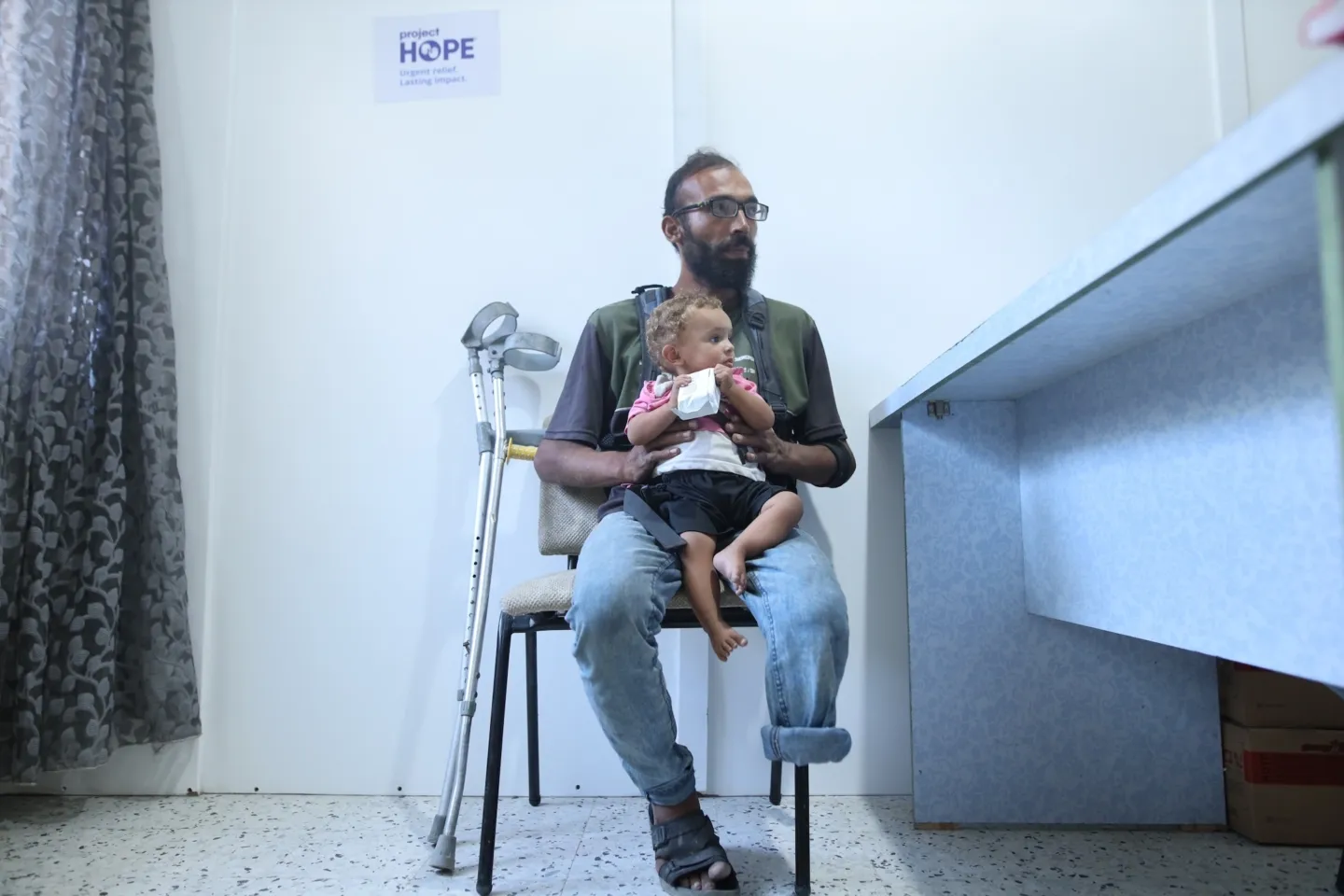 Man with amputated leg, holds young child in arms while speaking to medical staff in Gaza