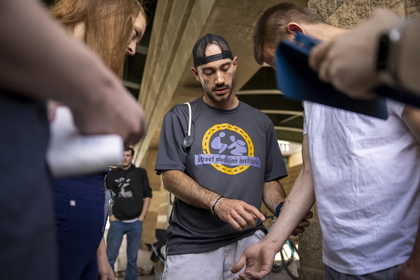Medical staffer with a backwards cap, "street medicine institute" t-shirt, and stethoscope checking arm of man on construction site.