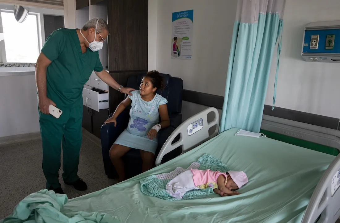 a doctor in scrubs checks a woman sitting in a chair while a newborn baby sleeps on a hospital bed